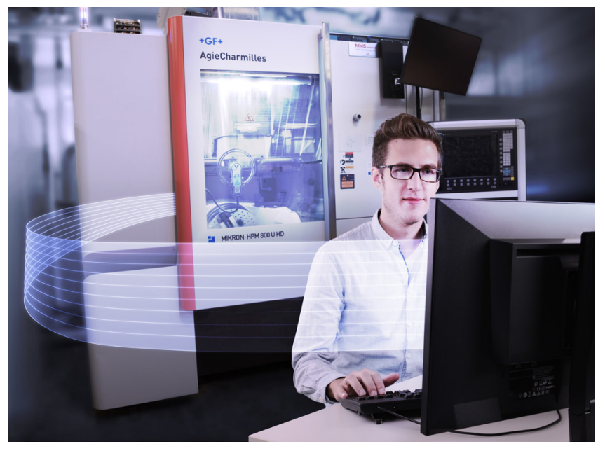 A person wearing a light-colored shirt is sitting at a desk with a computer, typing on a keyboard. In the background, there is a large, sophisticated machine labeled "Mikron HPM 800 U HD" with a transparent interface display showing a graphical representation of machinery, possibly for a manufacturing process. The setting is modern and sleek, with various monitors visible, including one on the right side displaying technical information. The overall color scheme includes cool tones such as grays and blues, and there are soft, flowing lines illustrated in white, suggesting a dynamic or digital element in the workspace.